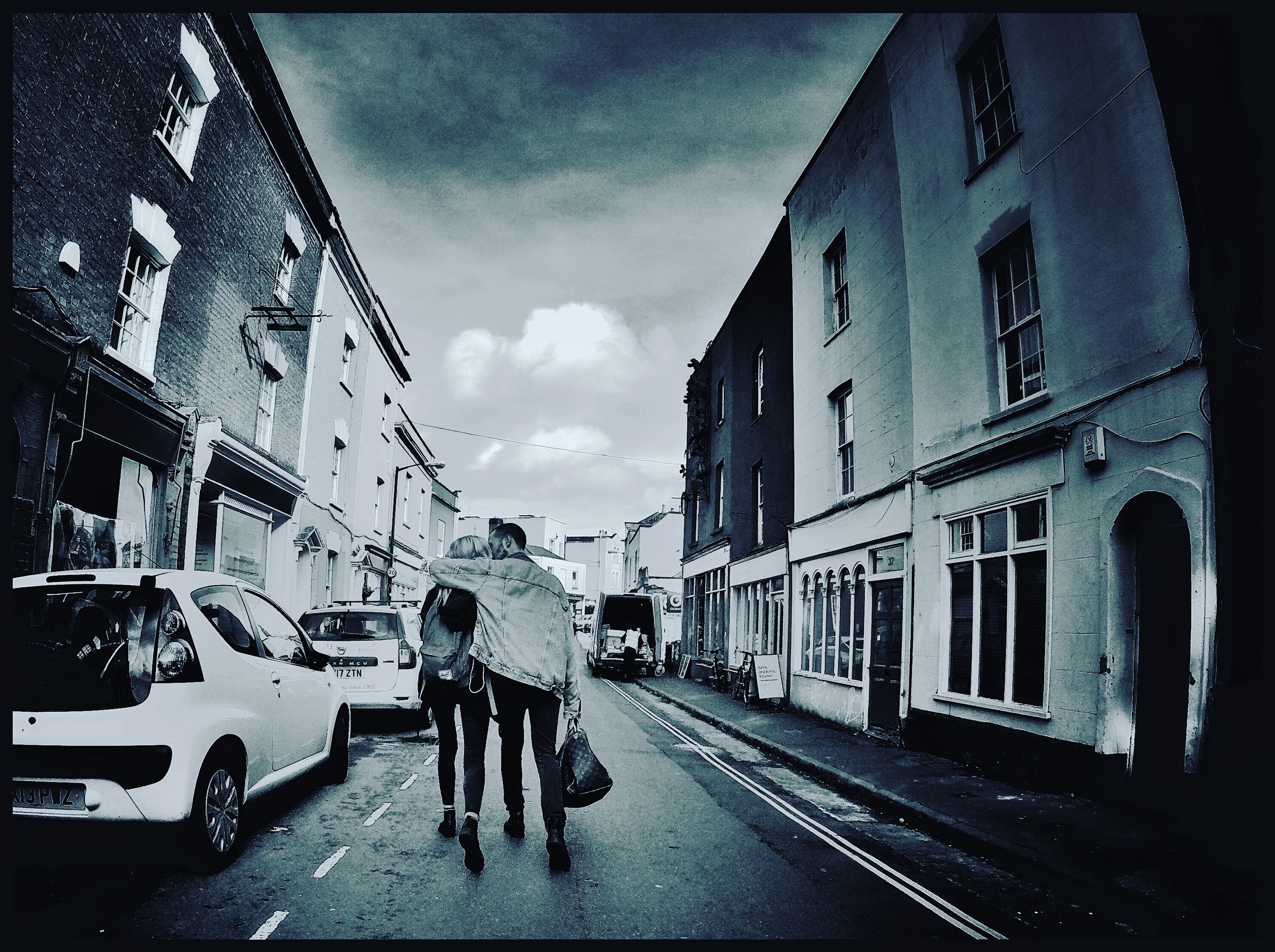 Two people walk down Picton Street in Bristol. One has their arm around the other. The photograph is desaturated.
